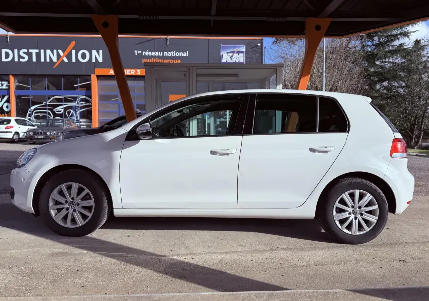 Profil côté gauche d'une Volkswagen Golf blanche 5 portes de 2012, stationnée devant un bâtiment commercial.