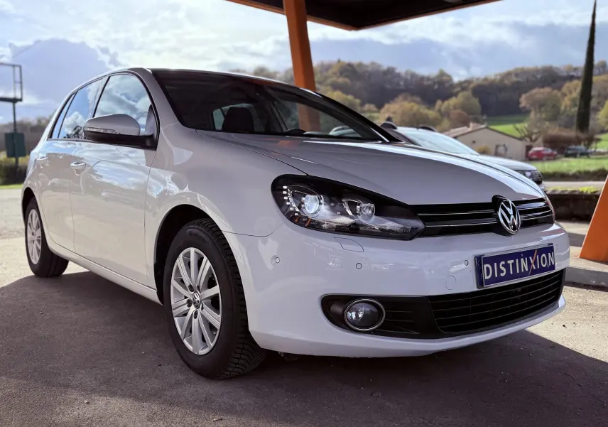 Volkswagen Golf blanche vue en 3/4 avant droit sous abri avec phares allumés et jantes argentées.