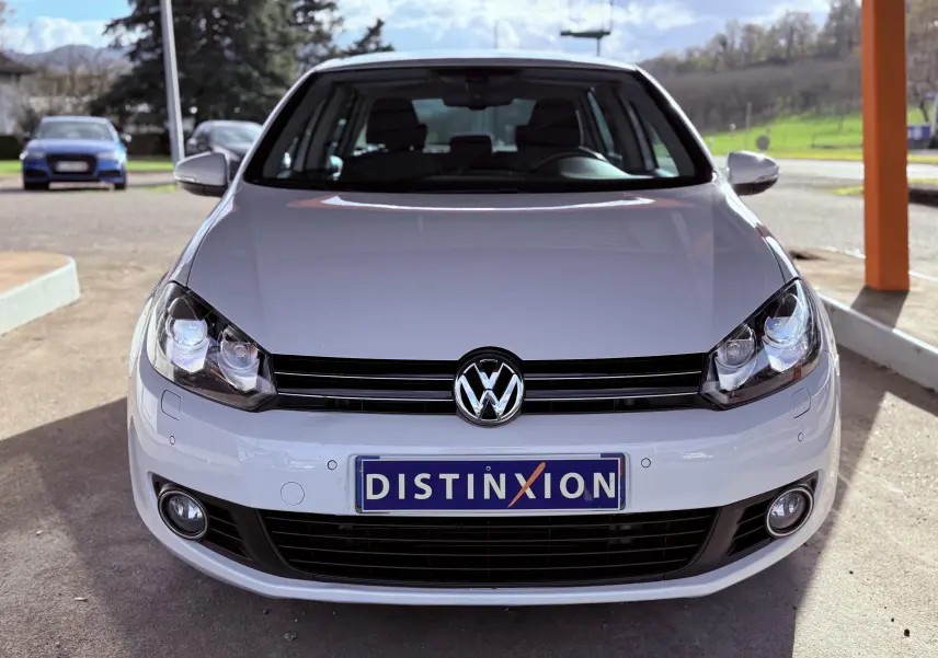 Vue frontale d'une Volkswagen Golf blanche de 2012 avec plaque Distinxion et phares allumés.