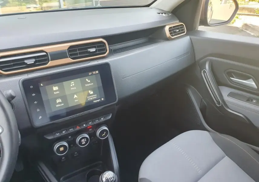 Vue intérieure côté passager du tableau de bord et console centrale du Dacia Duster gris urbain 2023 avec écran tactile et boîte manuelle.