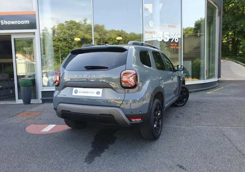 Vue 3/4 arrière droite d'un Dacia Duster gris urbain 2023 avec jantes noires et vitres surteintées, stationné devant un showroom.