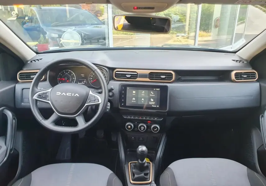 Vue intérieure frontale du tableau de bord du Dacia Duster 2023, avec volant, écran tactile et sellerie gris clair.