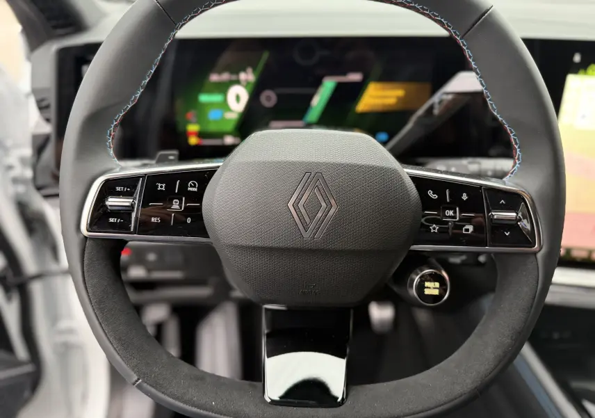Vue rapprochée du volant noir du Renault Austral Esprit Alpine, avec commandes intégrées et tableau de bord numérique flou en arrière-plan.