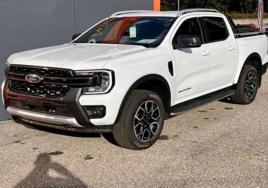 Ford Ranger Wildtrak blanc vu en 3/4 avant droit, avec jantes alu noires et barres de toit visibles.