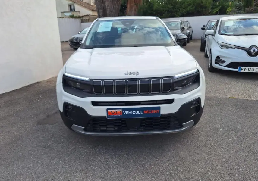 Vue frontale d'un Jeep Avenger blanc avec calandre noire et phares LED, stationné parmi d'autres véhicules.