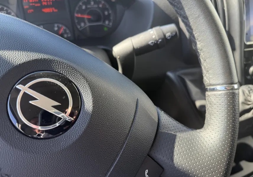Gros plan sur le volant en cuir perforé noir avec logo Opel, tableau de bord et commandes visibles en arrière-plan.