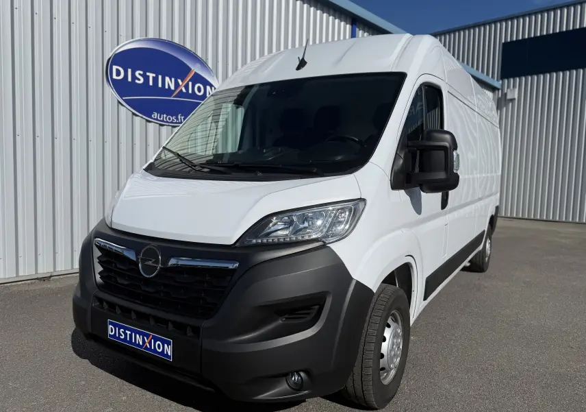 Vue 3/4 avant droit d’un Opel Movano Fourgon blanc 2023 avec pare-chocs noir et calandre chromée, stationné devant un bâtiment.