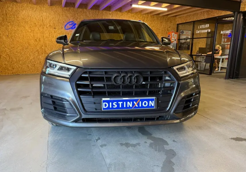 Vue avant d'un Audi Q5 Sportback gris 2019 avec calandre noire et plaques Distinxion en intérieur d'atelier.