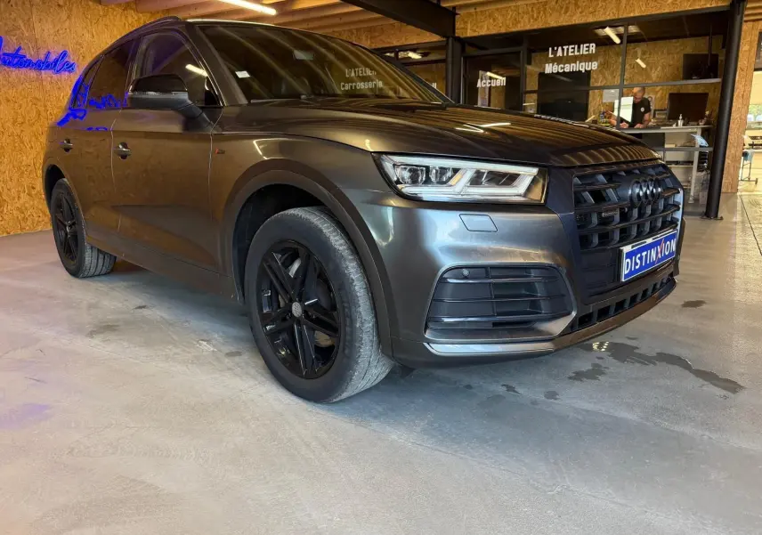 Audi Q5 Sportback gris en 3/4 avant droit, avec jantes noires et calandre marquée S-Line dans un garage.