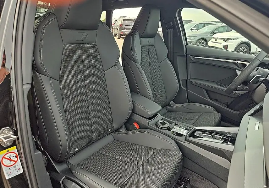 Vue intérieure côté avant gauche de l’Audi A3 Sportback 2026 avec sièges sport noirs et console centrale moderne.