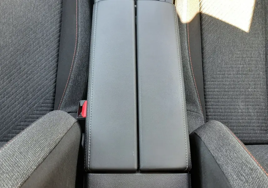 Vue plongeante sur l'accoudoir central noir et les sièges tissu gris foncé avec surpiqûres rouges du Peugeot 308 III Hybrid 2025.