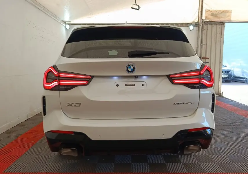 Vue arrière d’un BMW X3 xDrive30e blanc avec feux LED allumés et double sortie d’échappement dans un garage.