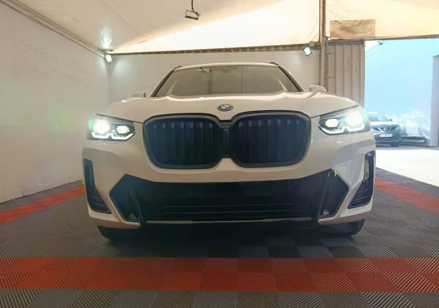 Vue frontale d'un BMW X3 blanc avec phares allumés et calandre noire dans un garage couvert.
