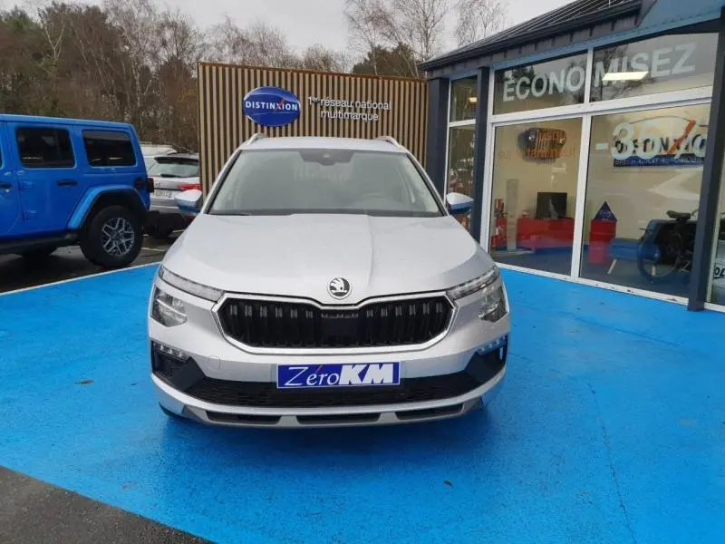 Vue avant d'un Skoda Kamiq gris diamant métallisé, avec calandre noire et phares LED, sur parking bleu devant un showroom.