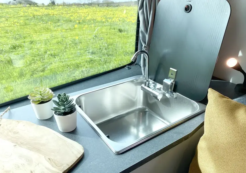 Gros plan intérieur sur l'évier inox avec robinet, plan de travail gris et fenêtre arrière du Renault Trafic Fourgon gris platine.