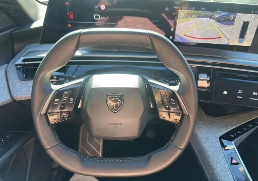 Vue rapprochée du volant cuir multifonctions Peugeot 5008 Hybrid 2025 avec tableau de bord numérique et écran caméra 360° affichés.