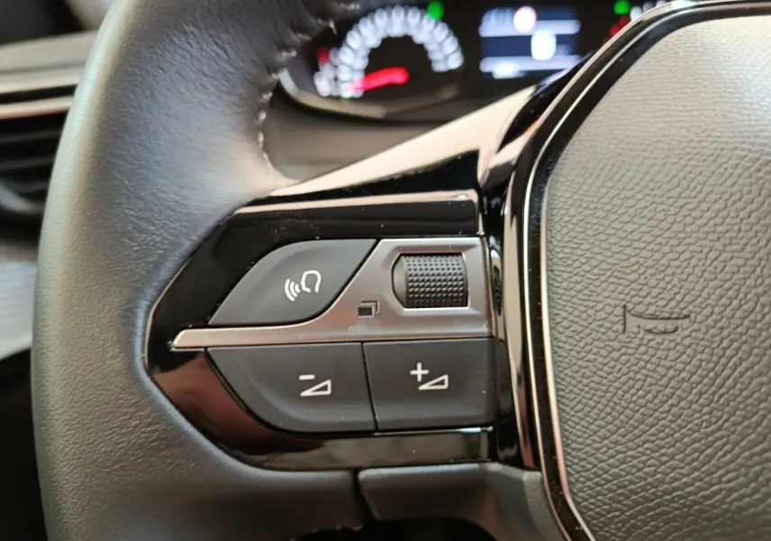 Gros plan sur les commandes gauche du volant cuir noir d’une Peugeot 208 blanche, avec tableau de bord flou en arrière-plan.
