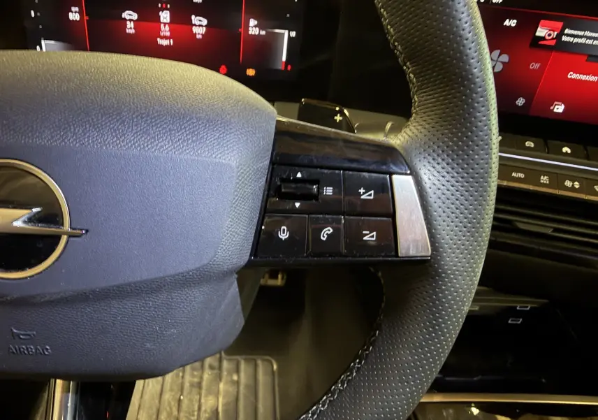 Gros plan sur le volant cuir perforé de l’Opel Astra 2024 avec commandes audio et palettes au volant, tableau de bord numérique en arrière-plan.