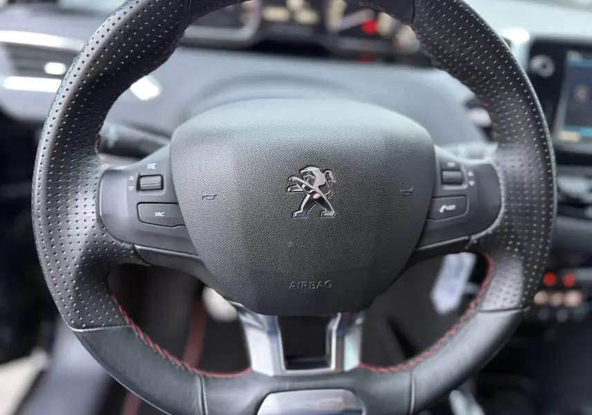 Gros plan sur le volant perforé avec surpiqûres rouges du Peugeot 2008 GT Line noir, tableau de bord flou en arrière-plan.