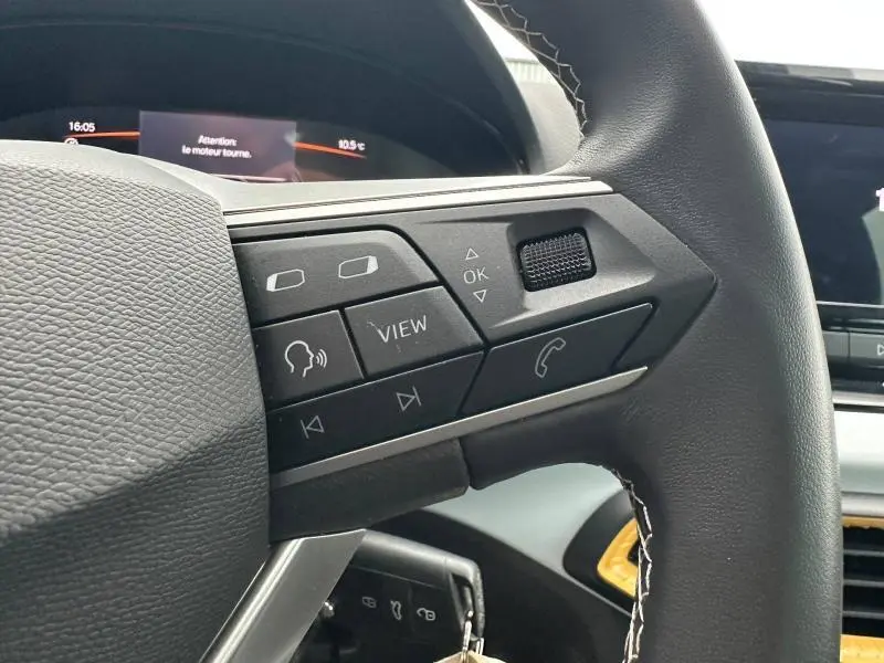 Gros plan sur les commandes au volant du SEAT Arona 2025, cuir noir avec surpiqûres blanches, affichage digital en arrière-plan.