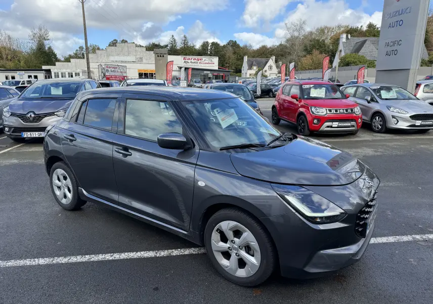Suzuki Swift 1.2 Hybrid Privilege gris vue 3/4 avant droit sur parking de concession avec panneaux Suzuki en arrière-plan.