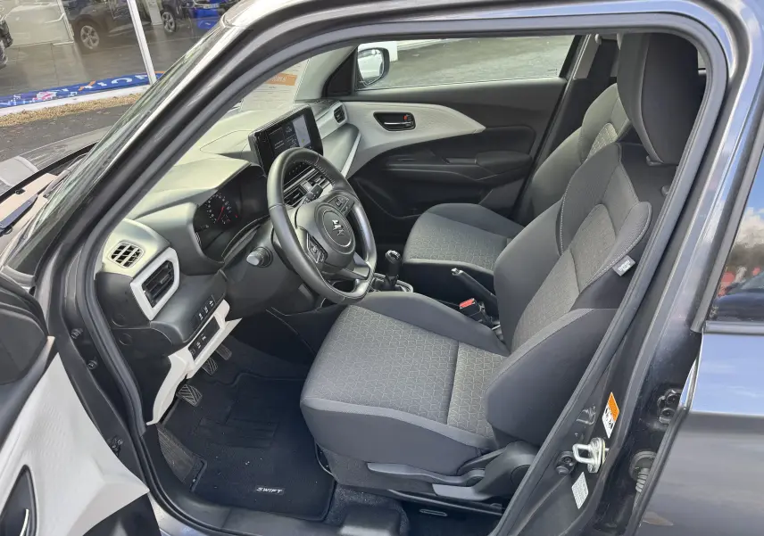 Vue intérieure côté conducteur de la Suzuki Swift 1.2 Hybrid Privilege gris, montrant sièges tissu et tableau de bord moderne.