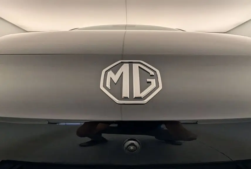 Gros plan sur le logo MG au centre d'une malle arrière Pebble Black Metal, avec caméra de recul visible en dessous.