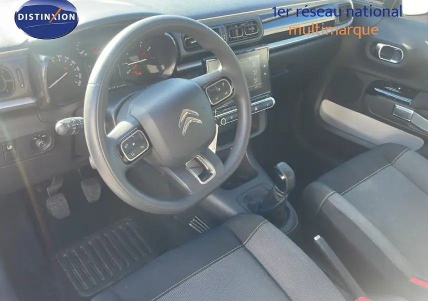Vue intérieure de face du poste de conduite d'une Citroën C3 blanche, volant multifonction et levier de vitesse manuel visibles.