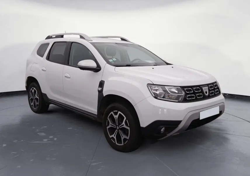 Dacia Duster blanc en 3/4 avant droit, avec calandre noire et jantes alliage, stationné sur un sol gravillonné.