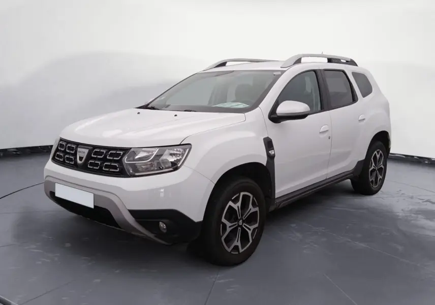 Dacia Duster blanc vue 3/4 avant droit sur parking, avec jantes alliage noires et toit barres de toit grises.