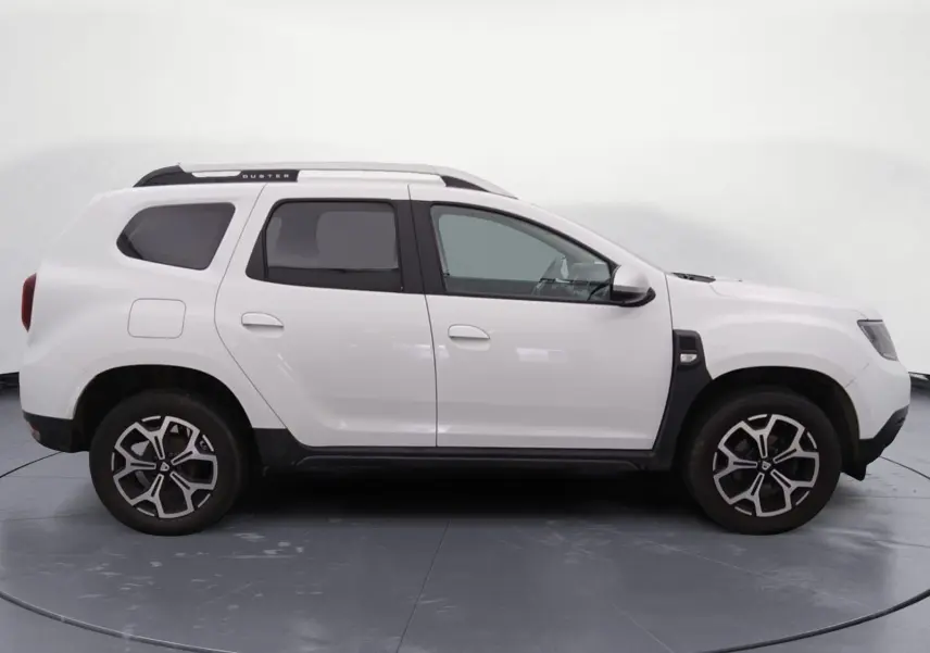 Profil droit d'un Dacia Duster blanc 2021 avec jantes alliage noires et toit noir, stationné sur un sol gravillonné.