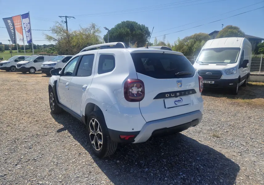 Vue 3/4 arrière droite d’un Dacia Duster blanc 2021 avec jantes alliage noires et toit ouvrant, stationné sur un terrain gravelé.