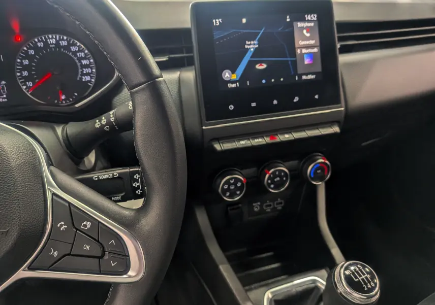 Vue intérieure centrée sur le volant et la console centrale avec écran tactile et levier de vitesse manuel de la Renault Clio V blanc glacier.