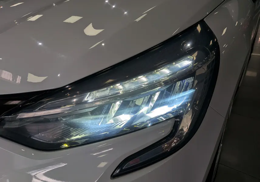 Gros plan sur le phare avant droit allumé d'une Renault Clio V Blanc Glacier, soulignant son design moderne et lumineux.