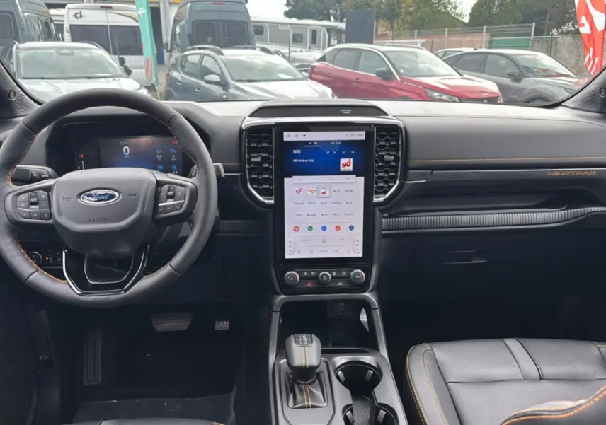 Vue intérieure avant du Ford Ranger Wildtrak Plus 2025 noir agate, avec volant cuir, écran tactile vertical et surpiqûres orange.