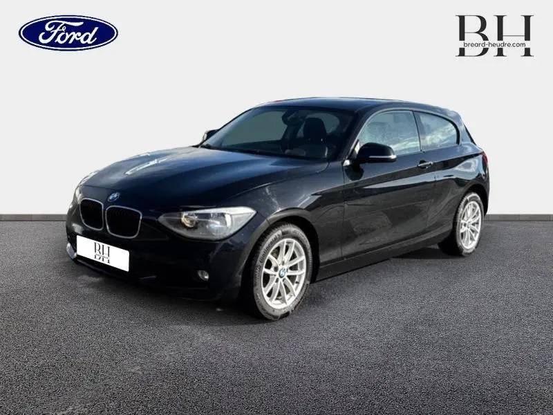 BMW Série 1 114i noire vue en 3/4 avant droit, avec jantes alu et lignes profilées visibles.