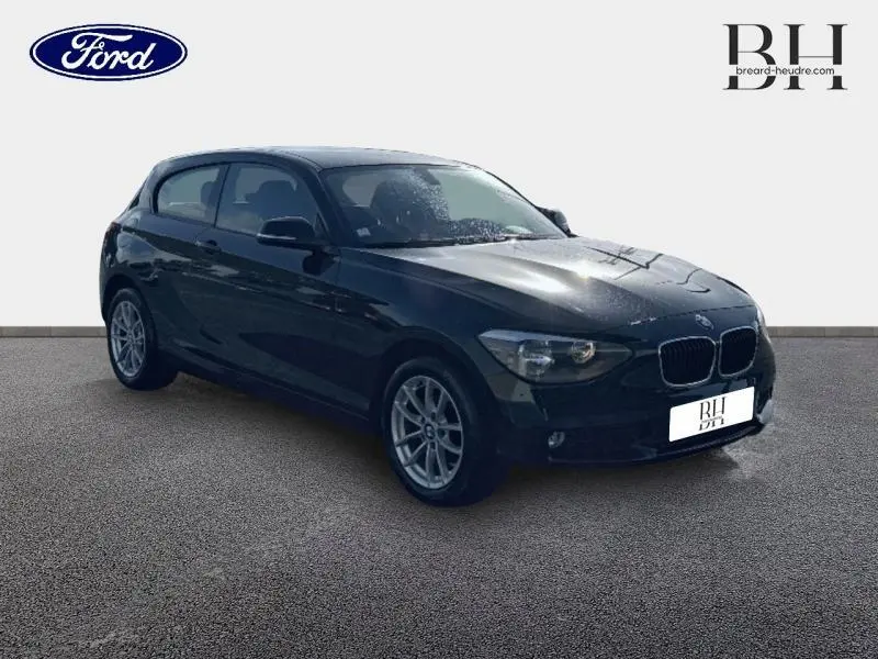 BMW Série 1 114i noire vue en 3/4 avant droit, avec jantes alu 16 pouces et phares halogènes allumés.