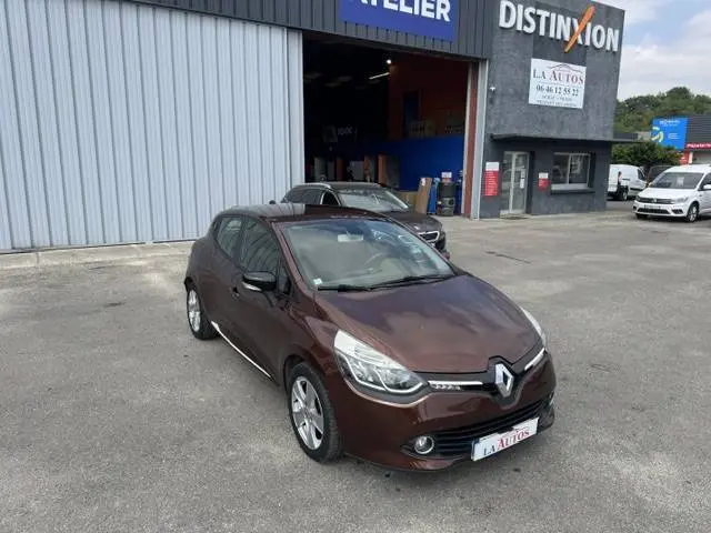Renault Clio IV marron vue 3/4 avant droit, avec barres de toit, stationnée devant un garage.