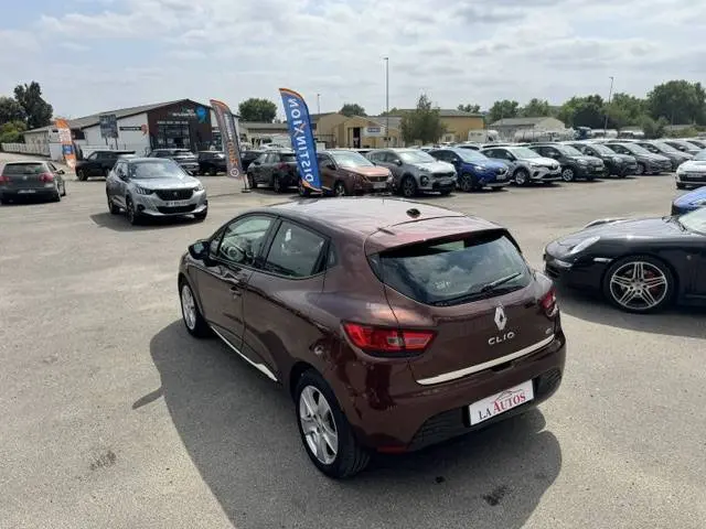 Vue 3/4 arrière droite d'une Renault Clio IV marron avec barres de toit, stationnée sur un parking extérieur.