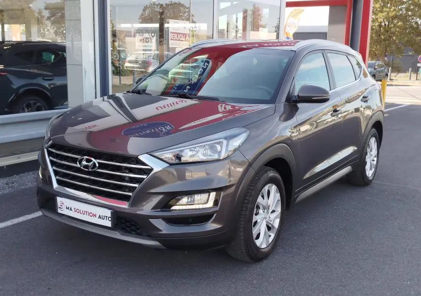 Vue 3/4 avant droite d'un Hyundai Tucson gris Micron 2019 avec calandre chromée et feux LED, stationné devant un showroom.