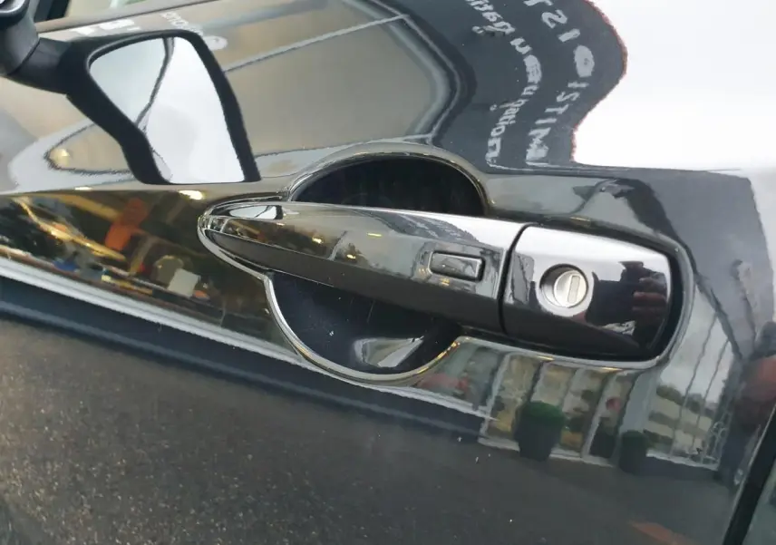 Poignée de porte noire côté gauche du Nissan Juke 2025 avec serrure et rétroviseur latéral réfléchissant.