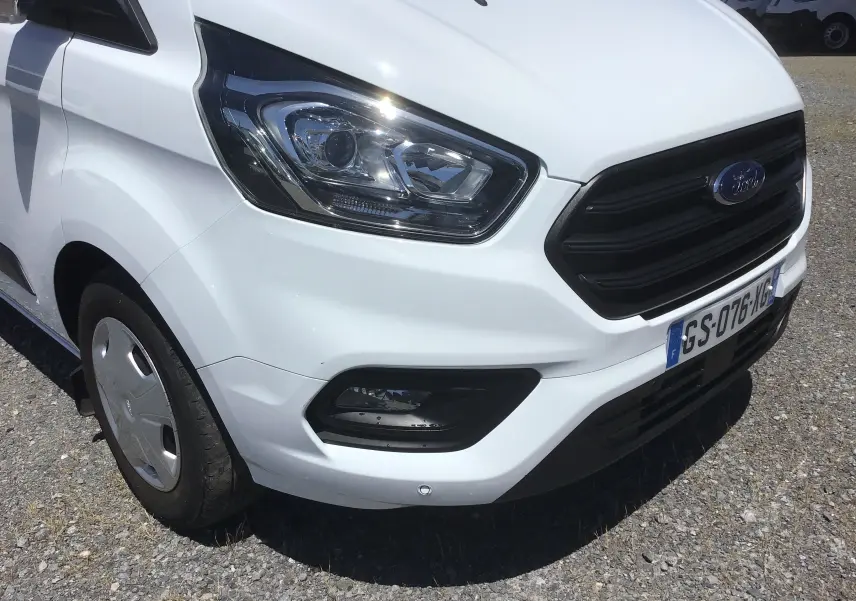 Vue 3/4 avant droit du Ford Transit Custom Kombi blanc, mettant en valeur le phare et la calandre noire.