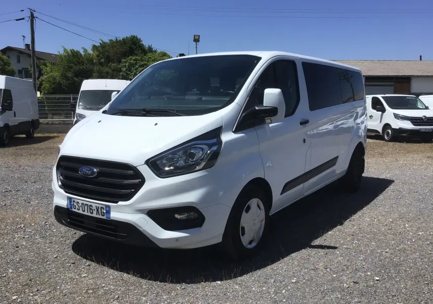 Ford Transit Custom Kombi blanc vu en 3/4 avant droit sur un parking avec plusieurs autres véhicules utilitaires.