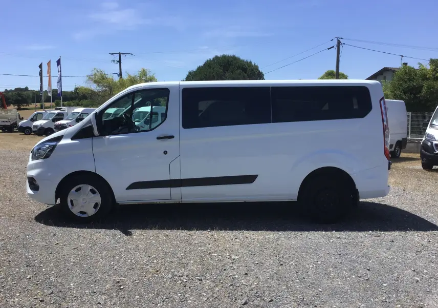 Vue de profil côté gauche d'un Ford Transit Custom Kombi blanc 2023 avec vitres arrière teintées et jantes différentes.