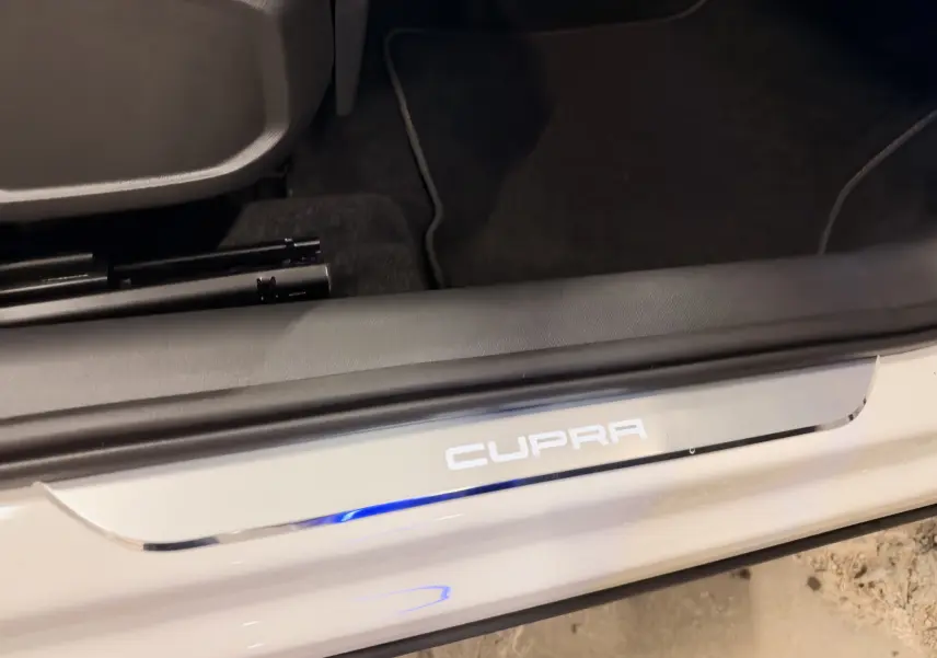 Gros plan sur le seuil de porte blanc avec logo CUPRA éclairé, intérieur noir visible au sol côté conducteur.