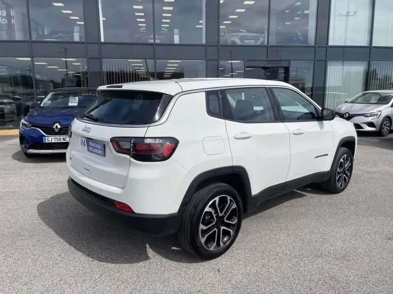 Jeep Compass blanc en 3/4 arrière droit, avec jantes alliage noires et vitres teintées, devant un showroom moderne.