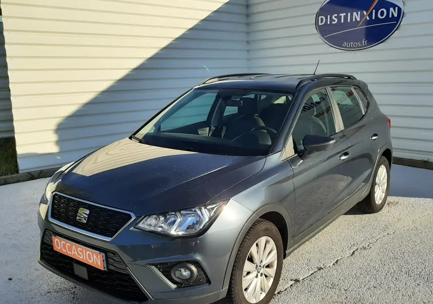 SEAT Arona gris magnetic avec toit noir, vue 3/4 avant droit, garée devant un mur blanc avec panneau Distinxion.