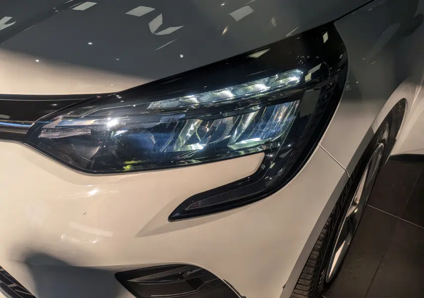 Gros plan sur le phare avant droit d'une Renault Clio V Blanc Glacier, soulignant les détails modernes du bloc optique.