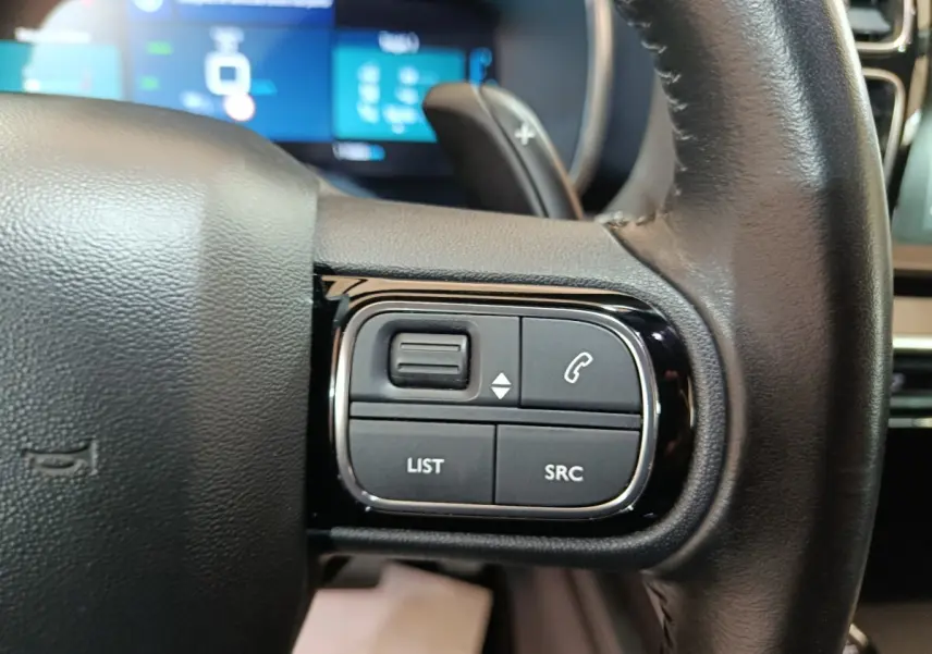 Gros plan sur les commandes gauche du volant noir du Citroën C5 Aircross Hybrid 225 ë-EAT8 avec affichage numérique en arrière-plan.