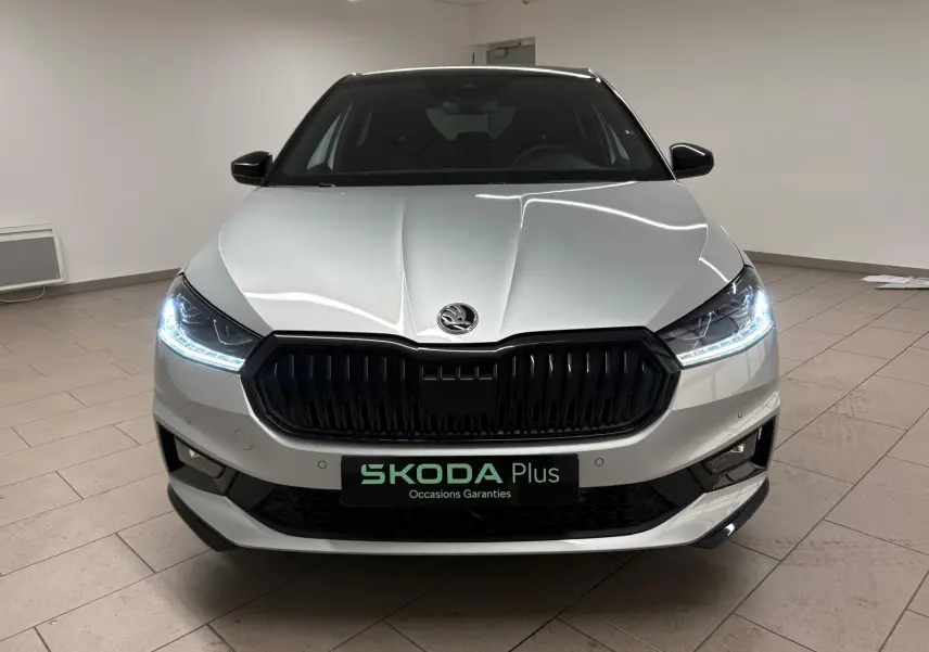 Vue frontale d'une Skoda Fabia 1.0 TSI 116 DSG7 Monte Carlo gris argent avec toit noir et phares allumés en intérieur.